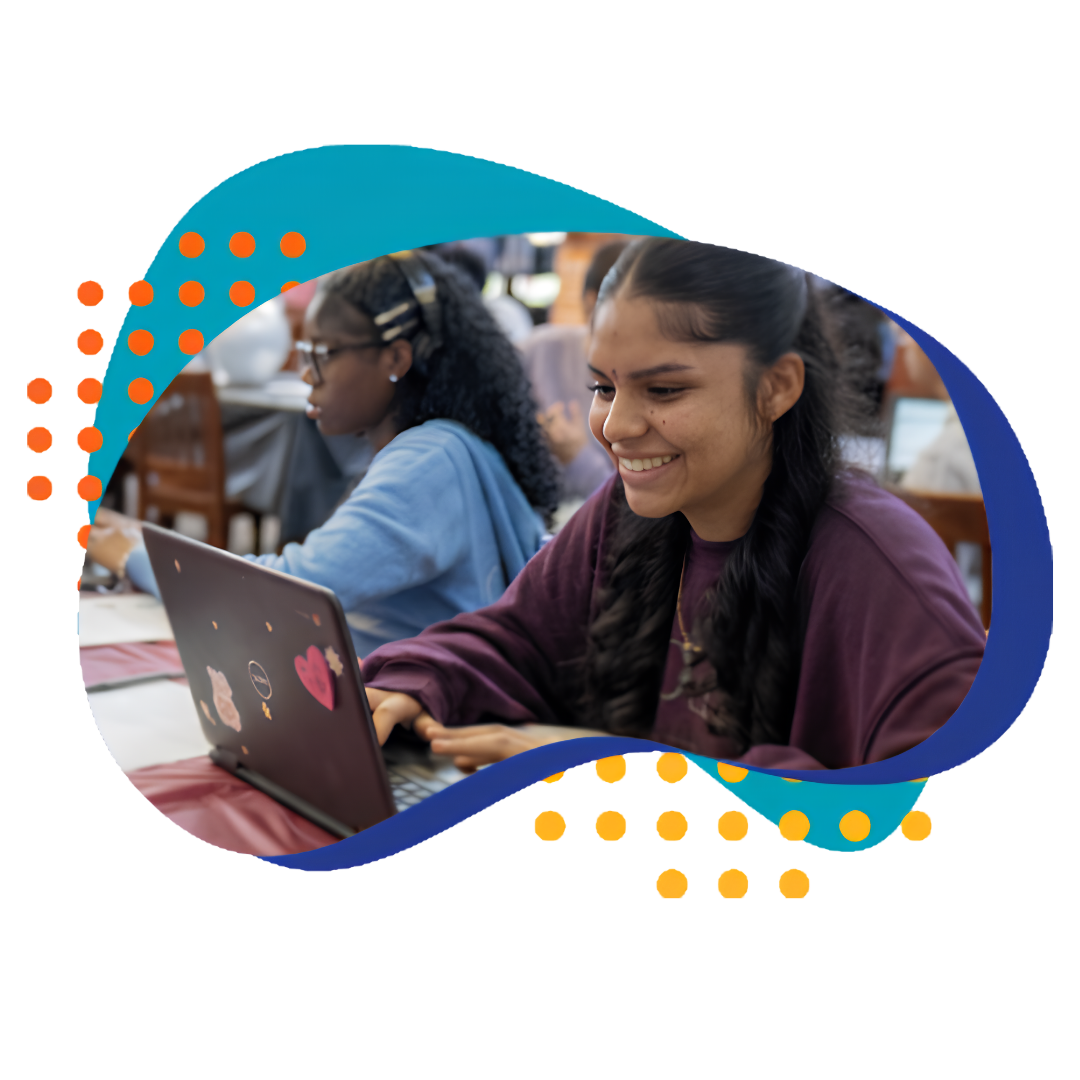 Two students study on laptops in a bright classroom, one smiling at the screen while the other works nearby with a blue sweater nearby and desks in the background.